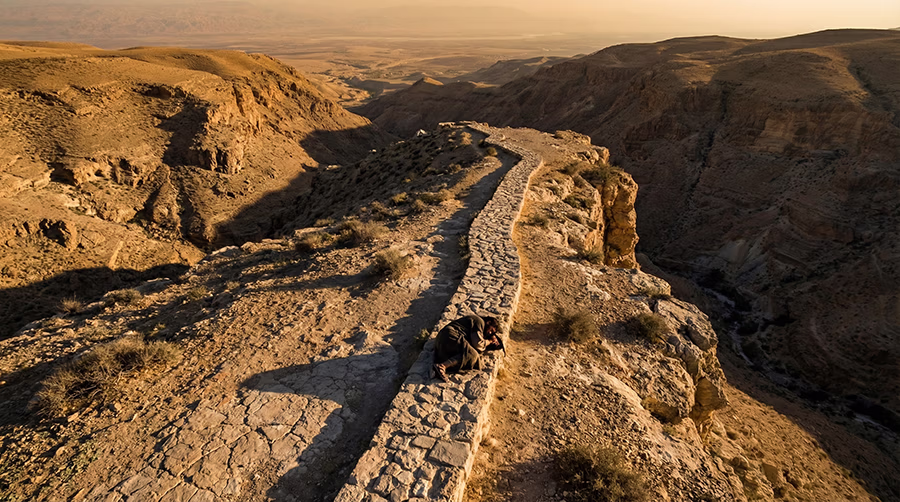 A wounded olive-skinned man in a torn bloodied linen tunic lies collapsed on the rocky Judean wilderness road between Jerusalem and Jericho